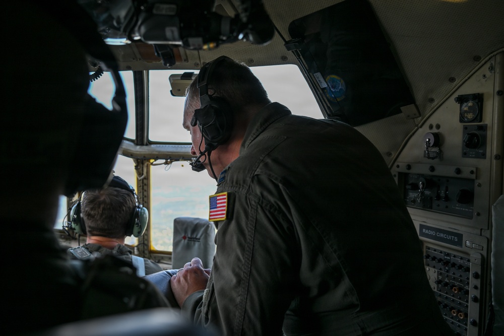 The image shows the interior of a military aircraft cockpit with at least two crew members visible. A prominent figure in the foreground wears an olive/dark green flight suit with a US flag patch on the shoulder and a communications headset. The crew appears to be in flight, with terrain visible through the cockpit windows.