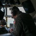 The image shows the interior of a military aircraft cockpit with at least two crew members visible. A prominent figure in the foreground wears an olive/dark green flight suit with a US flag patch on the shoulder and a communications headset. The crew appears to be in flight, with terrain visible through the cockpit windows.