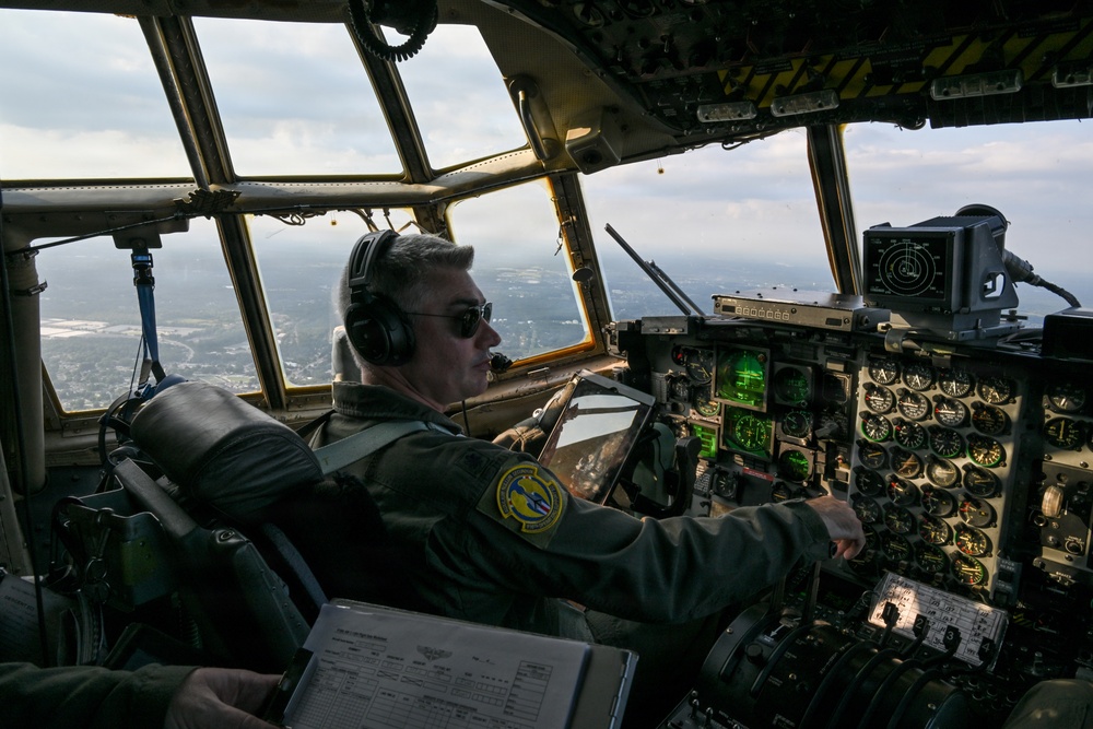 A military pilot is seated in the cockpit of a large military transport aircraft, actively operating the flight controls at altitude. The cockpit features an extensive array of analog and digital instruments, consistent with an older-generation military transport such as a C-130 Hercules or similar aircraft. The view through the large multi-pane windshield reveals a landscape below with partial cloud cover.