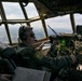 A military pilot is seated in the cockpit of a large military transport aircraft, actively operating the flight controls at altitude. The cockpit features an extensive array of analog and digital instruments, consistent with an older-generation military transport such as a C-130 Hercules or similar aircraft. The view through the large multi-pane windshield reveals a landscape below with partial cloud cover.