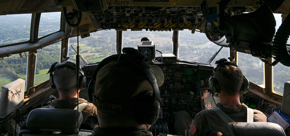 The image shows the interior of a military aircraft cockpit with three crew members visible from behind, flying over a green, rural landscape with scattered towns visible below. The cockpit features an extensive array of analog and digital instruments, controls, and overhead panels. The wide windshield provides a panoramic view of the terrain below, suggesting low-to-medium altitude flight.