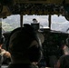 The image shows the interior of a military aircraft cockpit with three crew members visible from behind, flying over a green, rural landscape with scattered towns visible below. The cockpit features an extensive array of analog and digital instruments, controls, and overhead panels. The wide windshield provides a panoramic view of the terrain below, suggesting low-to-medium altitude flight.