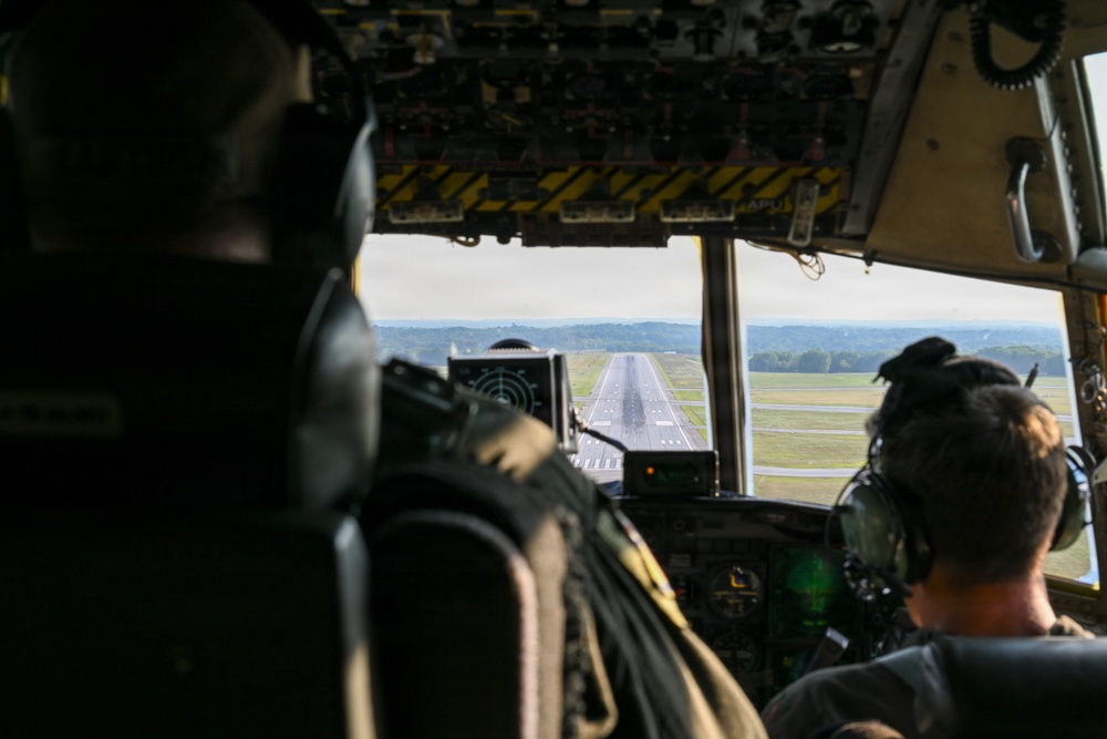 The image shows the interior of a military aircraft cockpit during approach to landing, with a runway clearly visible through the windshield. Two crew members wearing flight helmets and headsets are visible from behind, seated at their respective pilot and co-pilot positions. The cockpit features numerous overhead panels, instruments, and yellow-and-black hazard-striped panels typical of military transport aircraft.