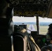 The image shows the interior of a military aircraft cockpit during approach to landing, with a runway clearly visible through the windshield. Two crew members wearing flight helmets and headsets are visible from behind, seated at their respective pilot and co-pilot positions. The cockpit features numerous overhead panels, instruments, and yellow-and-black hazard-striped panels typical of military transport aircraft.