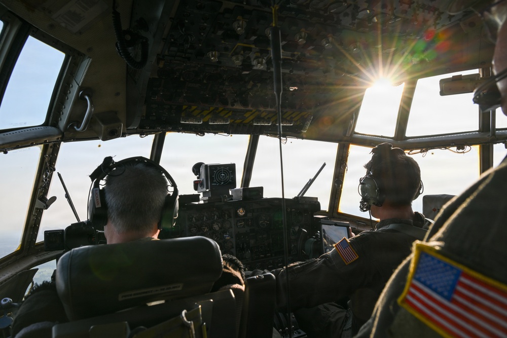 Two military aircrew members are seated in the cockpit of what appears to be a C-130 Hercules military transport aircraft in flight. Both personnel are wearing olive drab flight suits with American flag patches and aviation headsets. The image is taken from behind the crew, looking forward through the multi-panel cockpit windows with bright sunlight creating a lens flare effect.