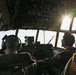 Two military aircrew members are seated in the cockpit of what appears to be a C-130 Hercules military transport aircraft in flight. Both personnel are wearing olive drab flight suits with American flag patches and aviation headsets. The image is taken from behind the crew, looking forward through the multi-panel cockpit windows with bright sunlight creating a lens flare effect.