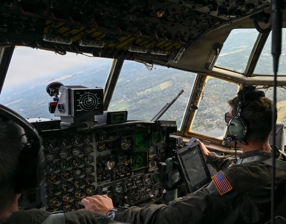 Two military aviators are visible in the cockpit of a large military transport aircraft, likely a C-130 Hercules, in flight over a green, wooded landscape with a visible river or lake below. The right-seat pilot is wearing a flight helmet with headset and has a US flag patch on his flight suit, and is referencing a tablet device. The cockpit features an extensive analog instrument panel with some modern digital upgrades.