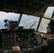 Two military aviators are visible in the cockpit of a large military transport aircraft, likely a C-130 Hercules, in flight over a green, wooded landscape with a visible river or lake below. The right-seat pilot is wearing a flight helmet with headset and has a US flag patch on his flight suit, and is referencing a tablet device. The cockpit features an extensive analog instrument panel with some modern digital upgrades.