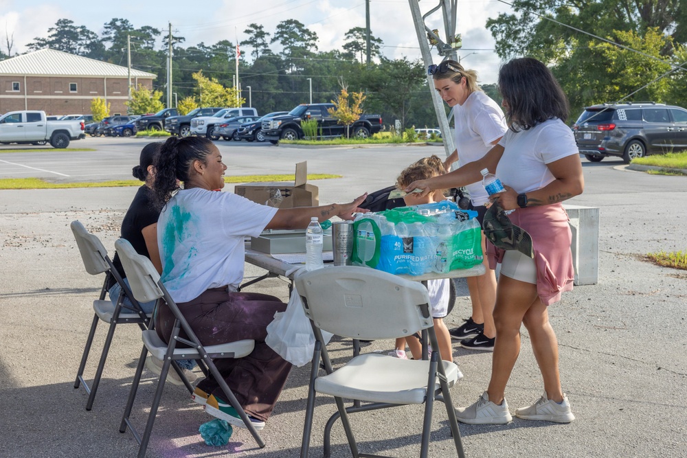 A group of people are gathered around a folding table in a parking lot, appearing to distribute or exchange water and supplies. Two individuals are seated at the table while others stand on the opposite side, with a case of purified water bottles prominently visible on the table. The setting suggests a community distribution event or relief effort.