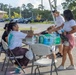 A group of people are gathered around a folding table in a parking lot, appearing to distribute or exchange water and supplies. Two individuals are seated at the table while others stand on the opposite side, with a case of purified water bottles prominently visible on the table. The setting suggests a community distribution event or relief effort.