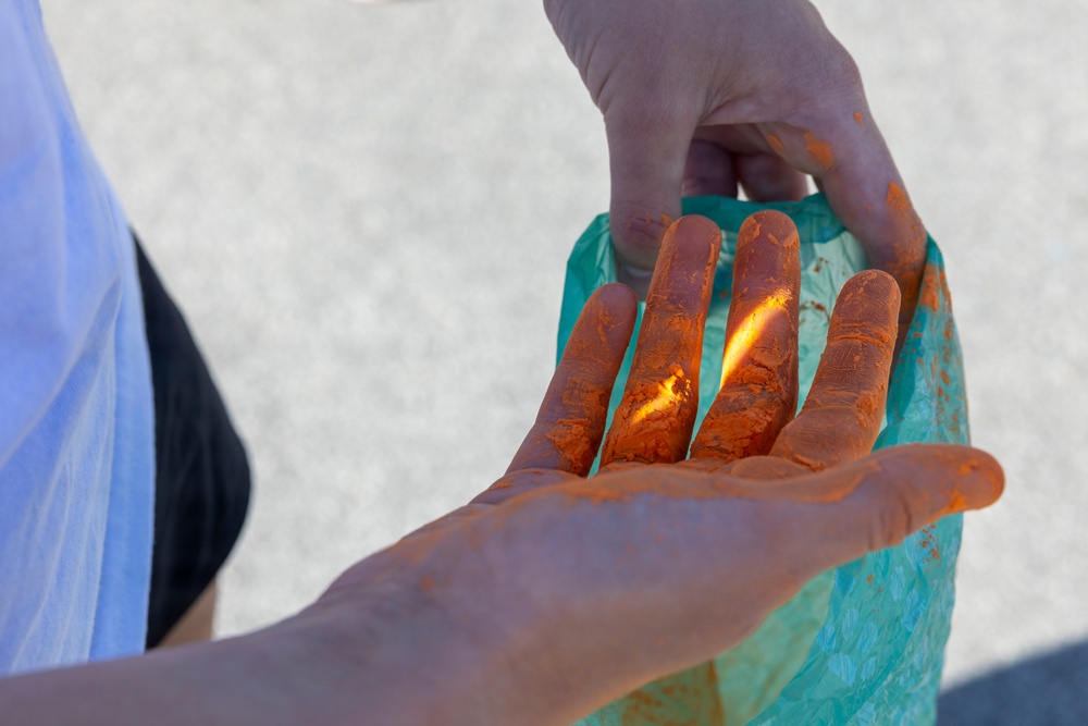 A close-up photograph showing a person's hands heavily coated with bright orange powder or paste-like substance. One hand is open and palm-up, displaying the orange coating across the fingers and palm, while the other hand holds a teal/turquoise plastic bag that is also stained with the orange material. The background is blurred and appears to be an outdoor paved surface.