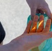 A close-up photograph showing a person's hands heavily coated with bright orange powder or paste-like substance. One hand is open and palm-up, displaying the orange coating across the fingers and palm, while the other hand holds a teal/turquoise plastic bag that is also stained with the orange material. The background is blurred and appears to be an outdoor paved surface.