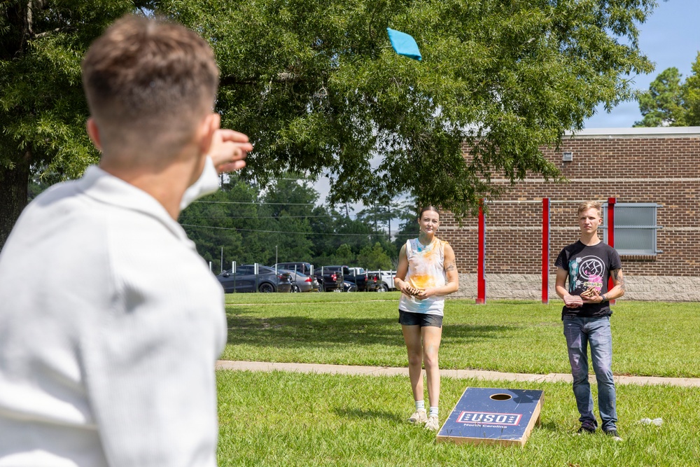 Three people are playing cornhole (bean bag toss) outdoors on a grassy area near a brick building. A person in a white hoodie in the foreground has just thrown a blue bean bag, which is visible in the air. Two players stand near a USO North Carolina branded cornhole board in the background.