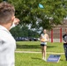 Three people are playing cornhole (bean bag toss) outdoors on a grassy area near a brick building. A person in a white hoodie in the foreground has just thrown a blue bean bag, which is visible in the air. Two players stand near a USO North Carolina branded cornhole board in the background.