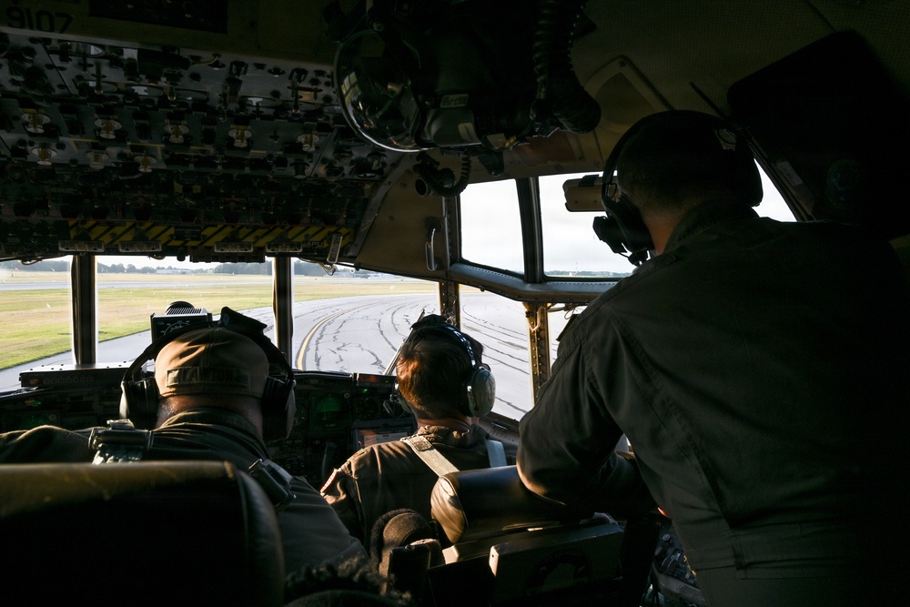 The image shows the interior of a military aircraft cockpit with three crew members seated and wearing aviation headsets. The crew appears to be on a runway or taxiway, visible through the large forward-facing cockpit windows. The aircraft appears to be a large military transport, likely a C-130 Hercules based on the cockpit configuration and overhead panel layout.