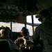 The image shows the interior of a military aircraft cockpit with three crew members seated and wearing aviation headsets. The crew appears to be on a runway or taxiway, visible through the large forward-facing cockpit windows. The aircraft appears to be a large military transport, likely a C-130 Hercules based on the cockpit configuration and overhead panel layout.