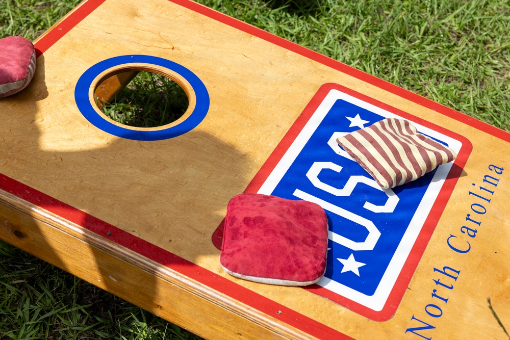 The image shows a cornhole board decorated with a 'USA' logo and 'North Carolina' text in blue cursive, placed on green grass. Several bean bags are visible on the board — two red/maroon solid ones and one striped red-and-white one. The board features a blue-ringed hole and red border trim, typical of regulation cornhole game equipment.