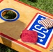 The image shows a cornhole board decorated with a 'USA' logo and 'North Carolina' text in blue cursive, placed on green grass. Several bean bags are visible on the board — two red/maroon solid ones and one striped red-and-white one. The board features a blue-ringed hole and red border trim, typical of regulation cornhole game equipment.