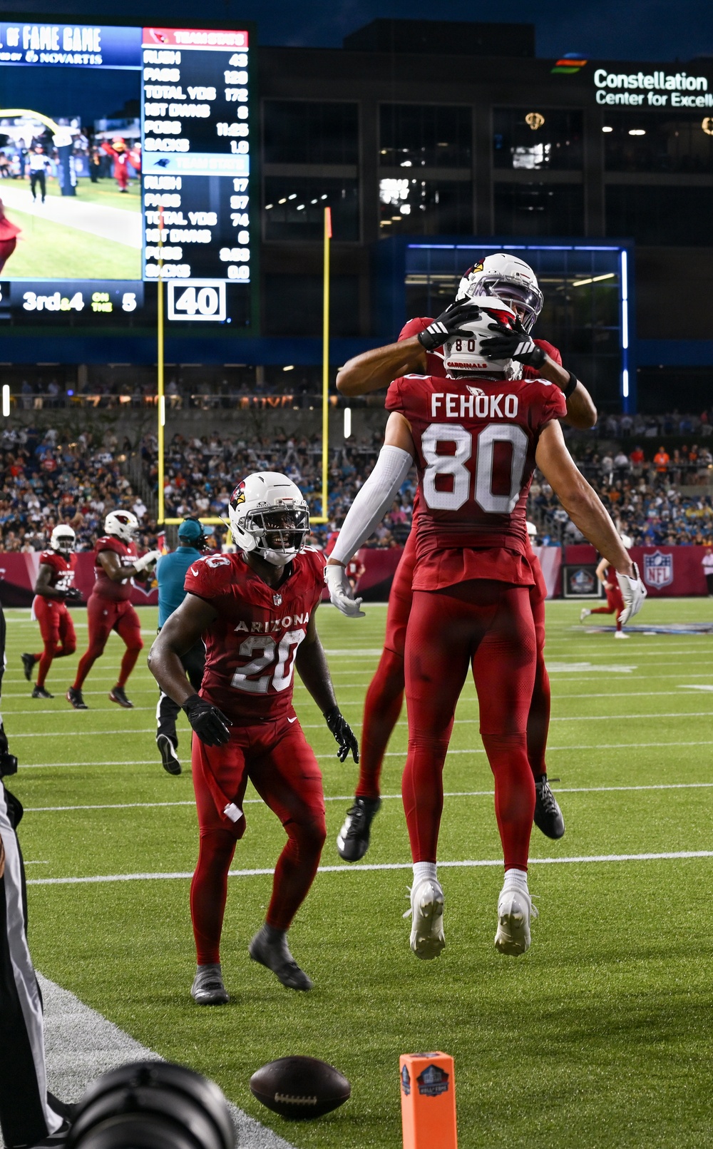 An NFL preseason game featuring the Arizona Cardinals celebrating what appears to be a touchdown near the end zone. Player #80 Fehoko is being lifted by a teammate in celebration, while #28 in an Arizona jersey looks on. The scoreboard in the background indicates a Hall of Fame Game, with game statistics visible on the display.