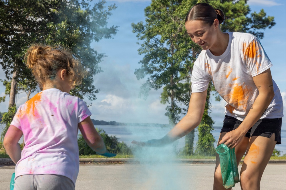 Two individuals — a young girl and an older female — are engaged in a color powder activity outdoors on a paved surface. The older female is leaning forward and releasing light blue powder from a bag toward the child, creating a visible cloud of color. Both participants have orange and other colored powder stains on their clothing, indicating prior color play.