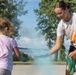 Two individuals — a young girl and an older female — are engaged in a color powder activity outdoors on a paved surface. The older female is leaning forward and releasing light blue powder from a bag toward the child, creating a visible cloud of color. Both participants have orange and other colored powder stains on their clothing, indicating prior color play.