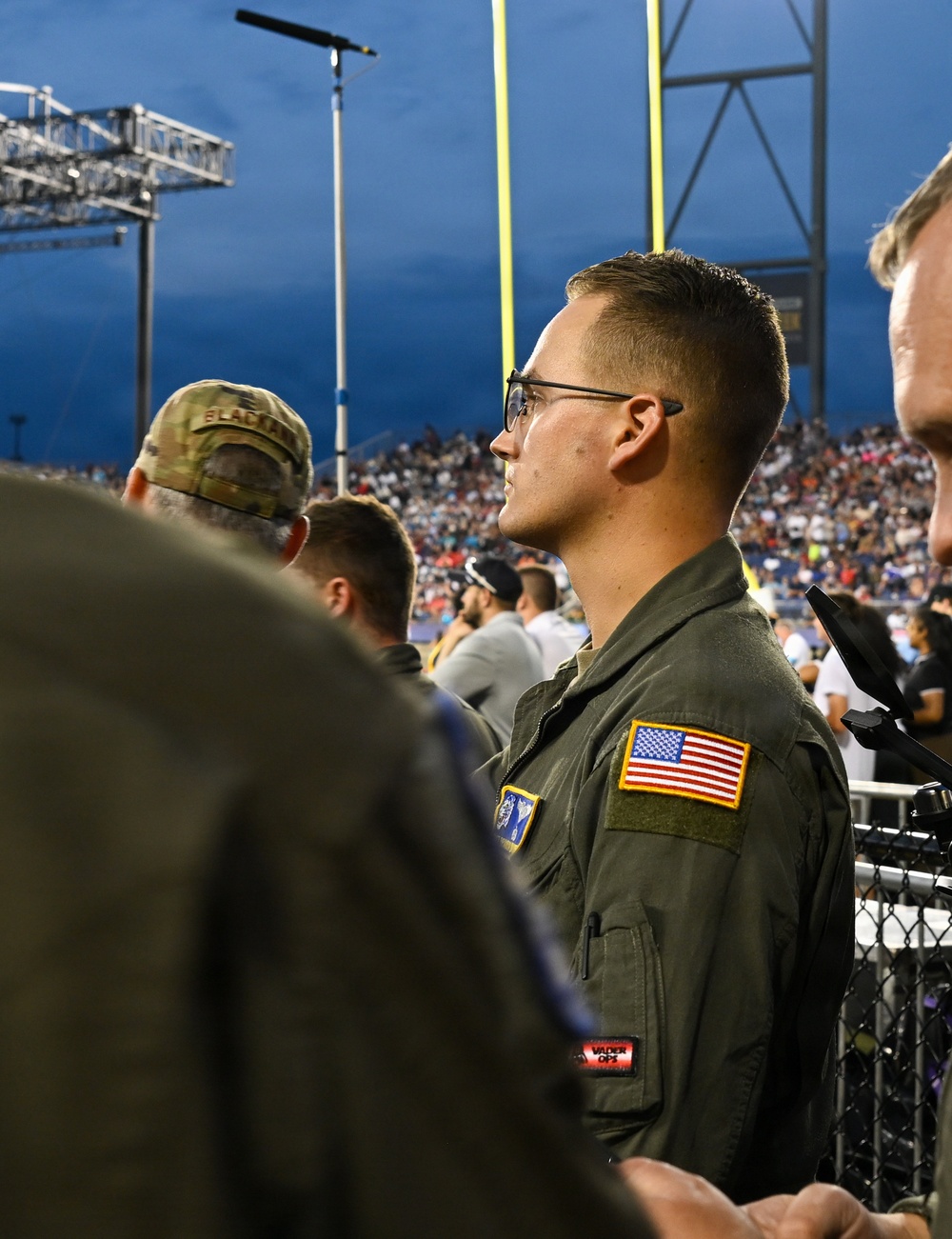 A young military service member wearing an olive drab flight suit with a US flag patch on the right shoulder stands at what appears to be an outdoor football stadium at night. He is facing forward attentively, possibly during a ceremony such as the national anthem. A crowd of spectators and yellow goal posts are visible in the background.