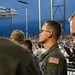 A young military service member wearing an olive drab flight suit with a US flag patch on the right shoulder stands at what appears to be an outdoor football stadium at night. He is facing forward attentively, possibly during a ceremony such as the national anthem. A crowd of spectators and yellow goal posts are visible in the background.