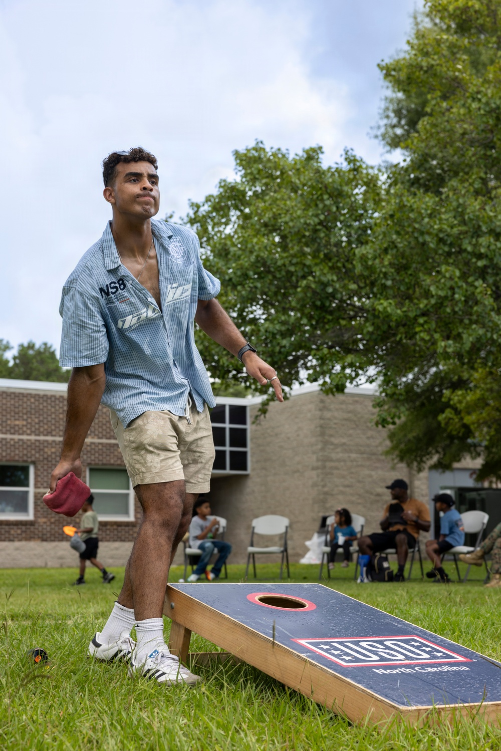 A young man is playing cornhole at an outdoor USO North Carolina event. He has just released a bean bag toss and is watching its trajectory while standing beside the cornhole board. In the background, a group of people are seated in folding chairs on a grassy lawn near brick buildings.