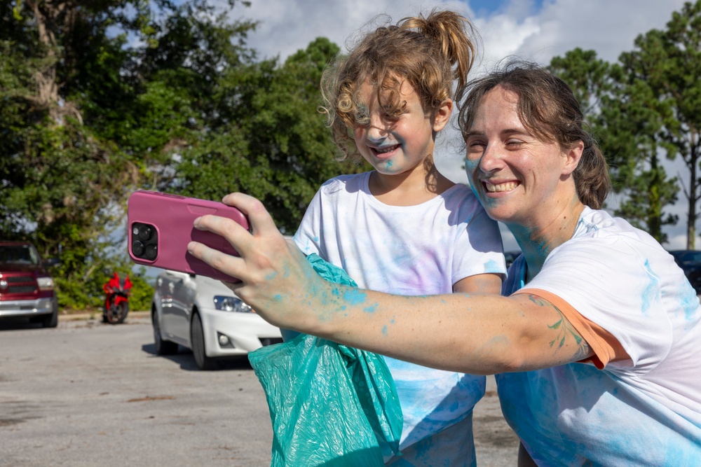 A woman and a young child are taking a selfie together in an outdoor parking lot setting. Both individuals are covered in blue powder/paint on their faces, arms, and white t-shirts, suggesting participation in a color run or similar powder-paint event. The woman is smiling broadly while holding a pink smartphone and a teal/turquoise bag.