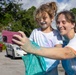 A woman and a young child are taking a selfie together in an outdoor parking lot setting. Both individuals are covered in blue powder/paint on their faces, arms, and white t-shirts, suggesting participation in a color run or similar powder-paint event. The woman is smiling broadly while holding a pink smartphone and a teal/turquoise bag.