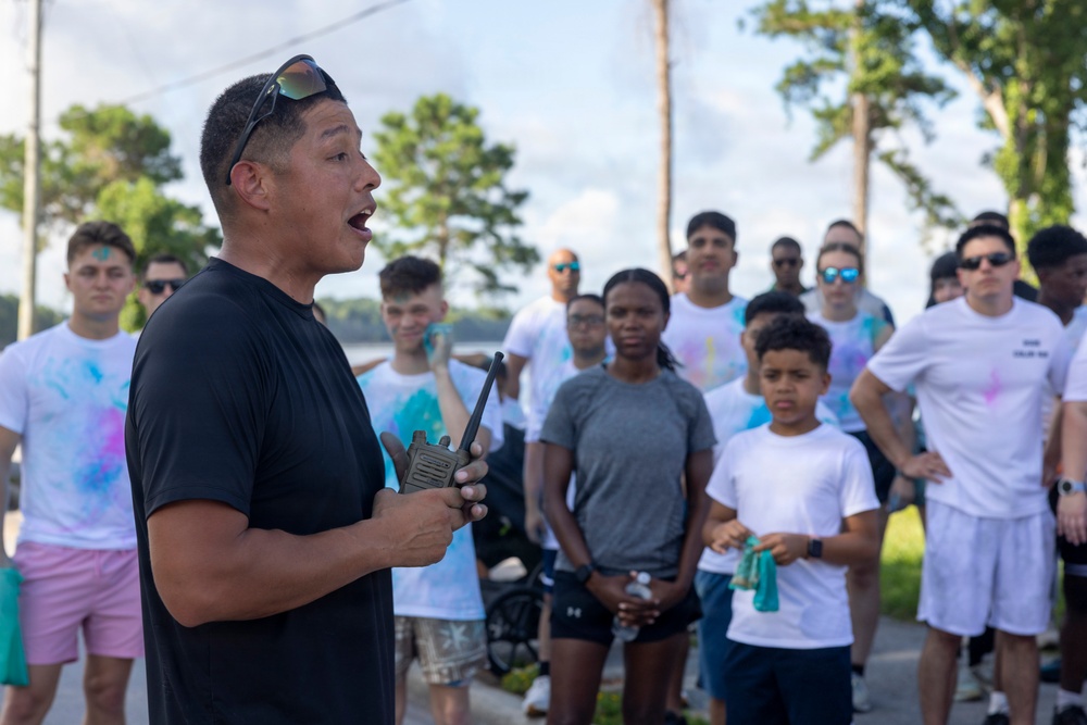 A man in a black t-shirt is addressing a large group of people outdoors while holding a walkie-talkie radio. The crowd, consisting of adults and at least one child, are wearing white t-shirts with visible colored powder stains, suggesting participation in a color run or similar event. The setting appears to be a paved outdoor area surrounded by trees.