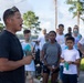 A man in a black t-shirt is addressing a large group of people outdoors while holding a walkie-talkie radio. The crowd, consisting of adults and at least one child, are wearing white t-shirts with visible colored powder stains, suggesting participation in a color run or similar event. The setting appears to be a paved outdoor area surrounded by trees.