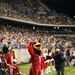 An Arizona Cardinals NFL mascot ('Big Red') poses on the sideline of a football field during a night game, giving a thumbs-up gesture. The scene includes cheerleaders, camera operators, and fans wearing Cardinals jerseys in a packed stadium. The setting appears to be a preseason or regular season NFL game with significant media presence on the sideline.