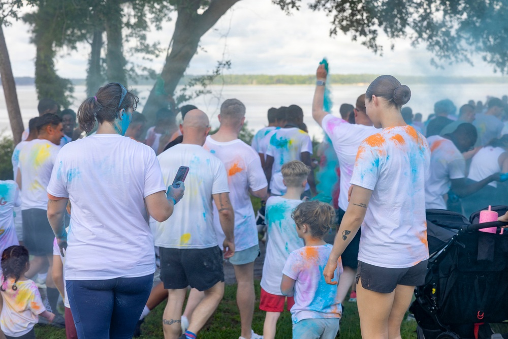 A group of participants wearing white t-shirts covered in colorful powder are gathered outdoors near a body of water, indicating a color run or Holi-style event. People of various ages, including children, are visible with paint splattered across their clothing in shades of blue, orange, yellow, and pink. One participant is raising their arm holding what appears to be a colored powder packet or smoke device.