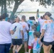 A group of participants wearing white t-shirts covered in colorful powder are gathered outdoors near a body of water, indicating a color run or Holi-style event. People of various ages, including children, are visible with paint splattered across their clothing in shades of blue, orange, yellow, and pink. One participant is raising their arm holding what appears to be a colored powder packet or smoke device.