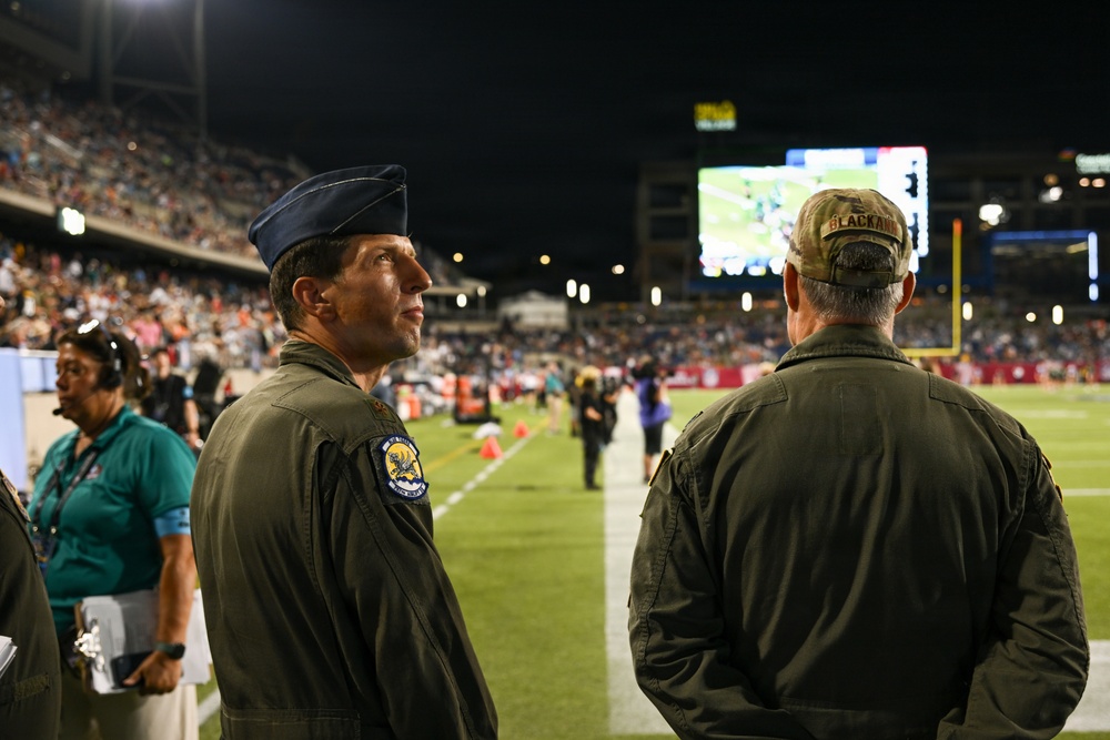 Two military personnel in olive drab flight suits are standing on the sideline of a nighttime football game in a stadium. The individual facing the camera wears a blue Air Force service cap and a unit patch on his left shoulder, while the other individual faces away wearing a camouflage cap with 'BLACKMAN' visible. The stadium is filled with spectators and a large video scoreboard is visible in the background.