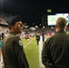 Two military personnel in olive drab flight suits are standing on the sideline of a nighttime football game in a stadium. The individual facing the camera wears a blue Air Force service cap and a unit patch on his left shoulder, while the other individual faces away wearing a camouflage cap with 'BLACKMAN' visible. The stadium is filled with spectators and a large video scoreboard is visible in the background.