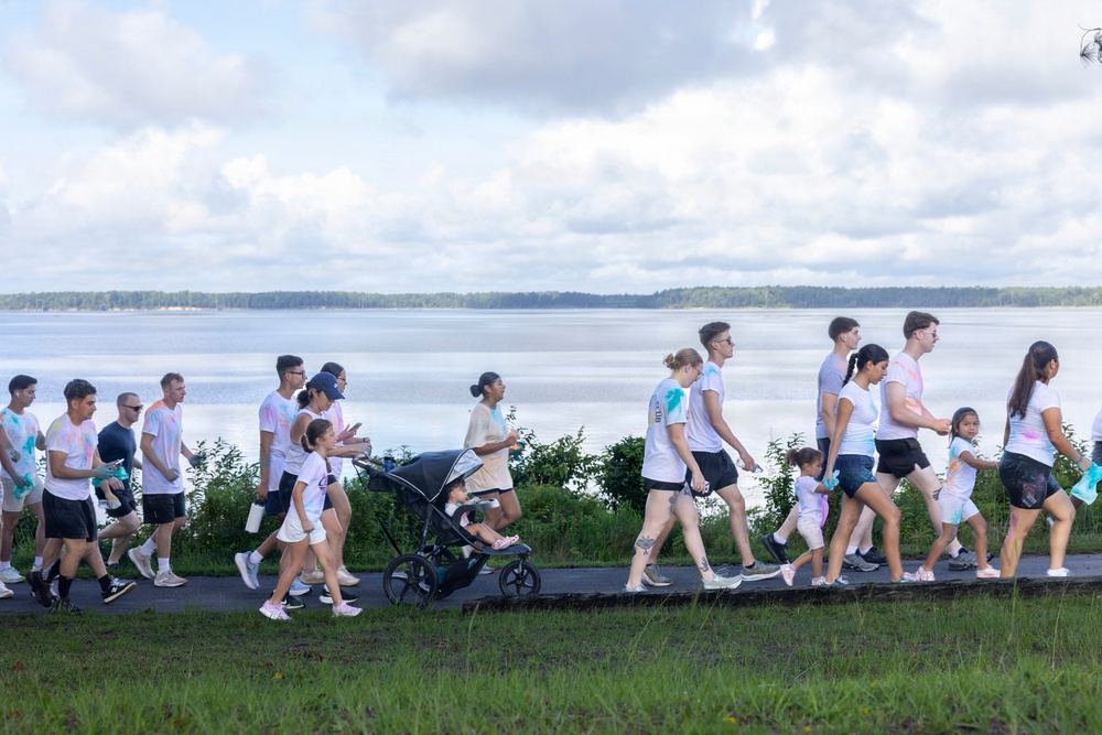 A group of people participating in what appears to be a color run or fun walk event along a paved path beside a large body of water. Most participants are wearing white t-shirts with visible colored powder stains (pink, blue, teal), suggesting they have passed through color stations. The group includes adults, children, and at least one infant in a stroller, indicating a family-friendly community event.