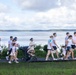 A group of people participating in what appears to be a color run or fun walk event along a paved path beside a large body of water. Most participants are wearing white t-shirts with visible colored powder stains (pink, blue, teal), suggesting they have passed through color stations. The group includes adults, children, and at least one infant in a stroller, indicating a family-friendly community event.