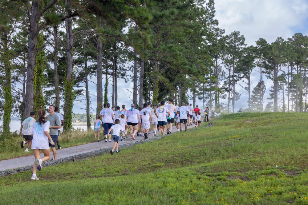 A large group of participants, predominantly wearing white t-shirts, are running or walking along a paved path lined with tall pine trees. The event appears to be a color run or fun run, as several shirts show traces of colored powder. The setting is an outdoor park or recreational area near a body of water visible in the background.