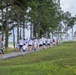 A large group of participants, predominantly wearing white t-shirts, are running or walking along a paved path lined with tall pine trees. The event appears to be a color run or fun run, as several shirts show traces of colored powder. The setting is an outdoor park or recreational area near a body of water visible in the background.