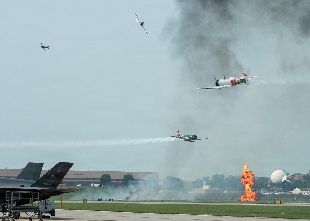 An airshow performance depicting a World War II Pacific theater reenactment, featuring vintage aircraft painted in Japanese Imperial markings conducting a simulated attack run. Pyrotechnic fire and smoke effects on the runway simulate bomb impacts, while multiple propeller-driven aircraft perform low passes. A modern F-35 fighter jet is visible on the static display ramp in the foreground.