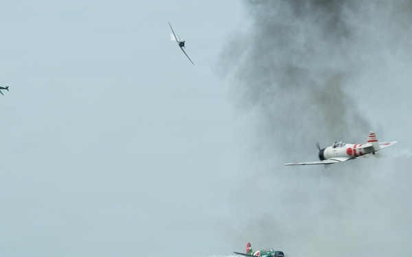 An airshow performance depicting a World War II Pacific theater reenactment, featuring vintage aircraft painted in Japanese Imperial markings conducting a simulated attack run. Pyrotechnic fire and smoke effects on the runway simulate bomb impacts, while multiple propeller-driven aircraft perform low passes. A modern F-35 fighter jet is visible on the static display ramp in the foreground.