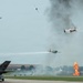 An airshow performance depicting a World War II Pacific theater reenactment, featuring vintage aircraft painted in Japanese Imperial markings conducting a simulated attack run. Pyrotechnic fire and smoke effects on the runway simulate bomb impacts, while multiple propeller-driven aircraft perform low passes. A modern F-35 fighter jet is visible on the static display ramp in the foreground.