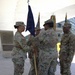 A military ceremony is taking place under a white architectural canopy structure, likely a change of command or guidon transfer ceremony. Four soldiers in U.S. Army OCP (Operational Camouflage Pattern) uniforms are participating, with one soldier passing or receiving a unit guidon/flag. A commemorative plaque is visible on the wall to the left, and a well-maintained military installation campus is visible in the background.