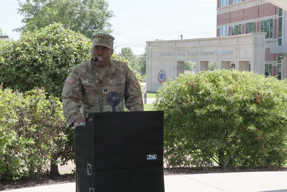 A U.S. Army Captain named Ford is speaking at an outdoor podium in front of the Combined Arms Support Command Headquarters building. The soldier is in full Army Combat Uniform (ACU) with a Captain's rank insignia visible on the chest. The event appears to be a formal military ceremony or address taking place on a sunny day.