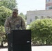 A U.S. Army Captain named Ford is speaking at an outdoor podium in front of the Combined Arms Support Command Headquarters building. The soldier is in full Army Combat Uniform (ACU) with a Captain's rank insignia visible on the chest. The event appears to be a formal military ceremony or address taking place on a sunny day.
