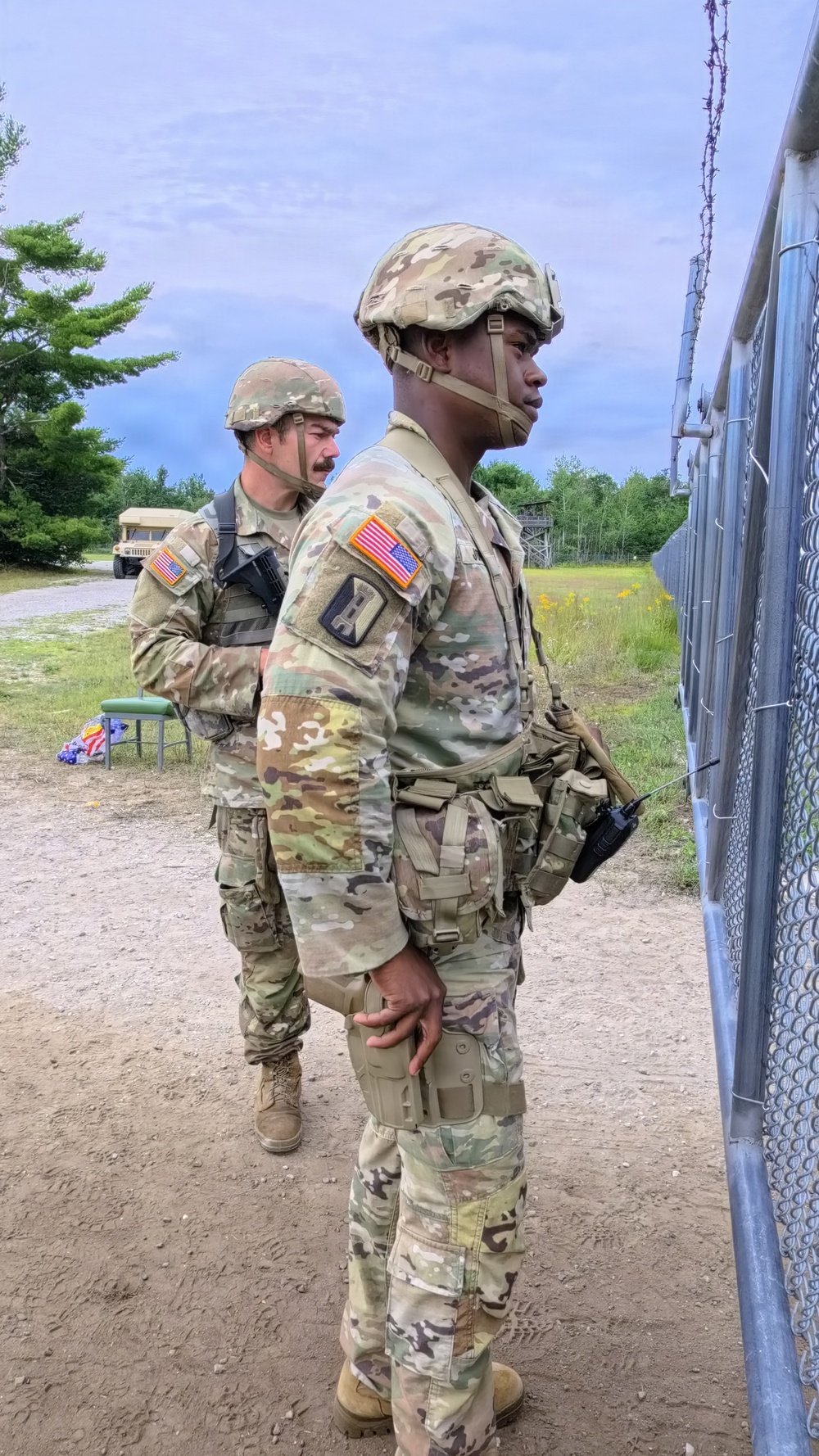 Two U.S. Army soldiers in Operational Camouflage Pattern (OCP) uniforms stand near a chain-link fence topped with barbed wire at what appears to be a military training installation. The foreground soldier wears a combat helmet, body armor, and carries a radio, with a Captain's rank insignia visible on the shoulder. A military vehicle (HMMWV/Humvee) is visible in the background.