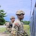 Two U.S. Army soldiers in Operational Camouflage Pattern (OCP) uniforms stand near a chain-link fence topped with barbed wire at what appears to be a military training installation. The foreground soldier wears a combat helmet, body armor, and carries a radio, with a Captain's rank insignia visible on the shoulder. A military vehicle (HMMWV/Humvee) is visible in the background.