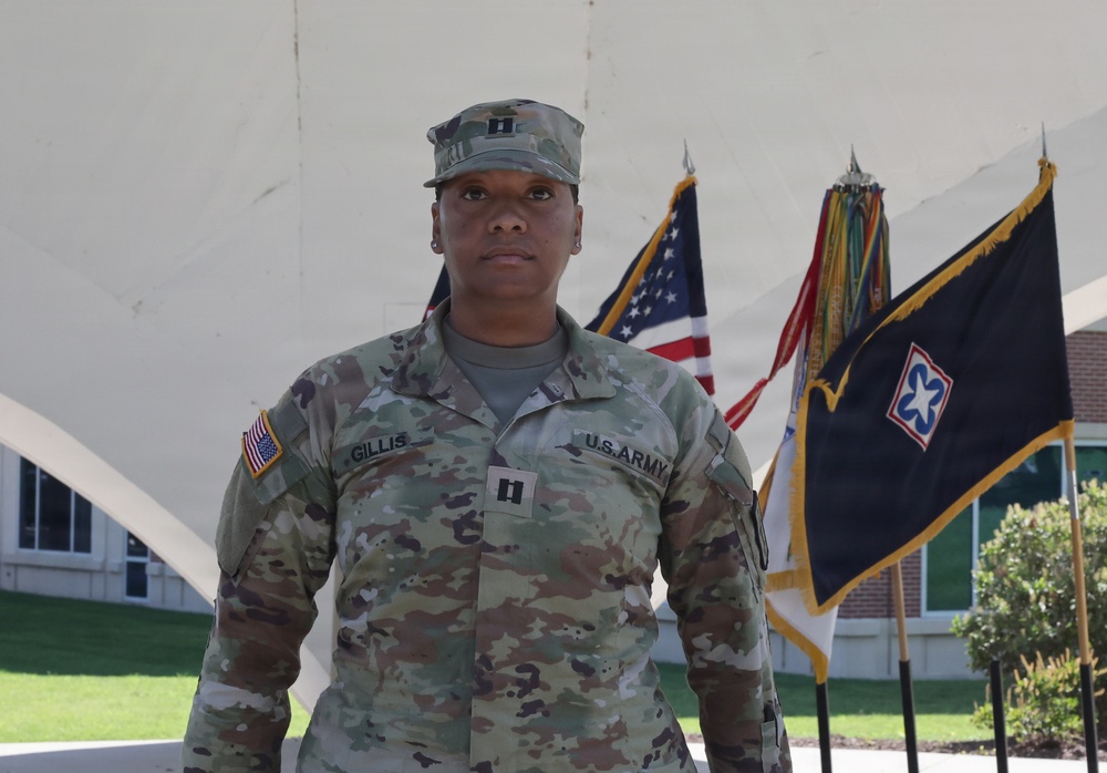 A U.S. Army Captain named Gillis stands at attention in Army Combat Uniform (ACU/OCP pattern) in front of a white architectural structure. The officer displays Captain rank insignia (two silver bars) on both the patrol cap and chest. Multiple military flags, including the U.S. flag and what appears to be a unit guidon with gold fringe, are visible in the background.